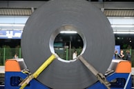 A large roll of coiled metal is secured with yellow straps and placed on a blue stand. The setting appears to be a train station platform with a few people in the background. Overhead lights brighten the scene, and there are informational signs visible near the top.