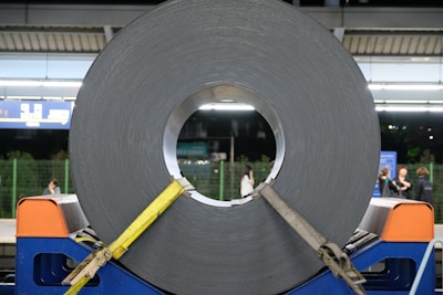 A large roll of coiled metal is secured with yellow straps and placed on a blue stand. The setting appears to be a train station platform with a few people in the background. Overhead lights brighten the scene, and there are informational signs visible near the top.