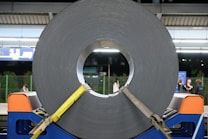 A large roll of coiled metal is secured with yellow straps and placed on a blue stand. The setting appears to be a train station platform with a few people in the background. Overhead lights brighten the scene, and there are informational signs visible near the top.