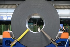 A large roll of coiled metal is secured with yellow straps and placed on a blue stand. The setting appears to be a train station platform with a few people in the background. Overhead lights brighten the scene, and there are informational signs visible near the top.