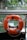 An orange lifebuoy with black text is attached to a railing, overlooking a calm body of water. There are green plants and rocks in the background, suggesting a natural setting.