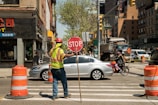 A construction worker wearing a reflective vest and a hard hat is holding a stop sign to manage traffic at an intersection in an urban area. The scene includes several vehicles, such as a silver car and a bicycle with a cyclist waiting nearby. Orange traffic cones are set up along the crosswalk, and buildings line the street with some storefronts and signs visible.