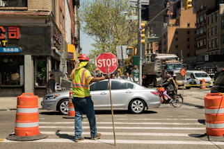 Industrial safety worker wearing reflective vest and helmet, holding a clipboard near traffic cones on a construction site.