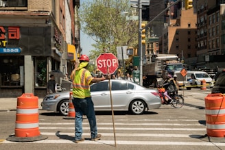 A construction worker wearing a reflective vest and a hard hat is holding a stop sign to manage traffic at an intersection in an urban area. The scene includes several vehicles, such as a silver car and a bicycle with a cyclist waiting nearby. Orange traffic cones are set up along the crosswalk, and buildings line the street with some storefronts and signs visible.