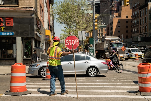 A construction worker wearing a reflective vest and a hard hat is holding a stop sign to manage traffic at an intersection in an urban area. The scene includes several vehicles, such as a silver car and a bicycle with a cyclist waiting nearby. Orange traffic cones are set up along the crosswalk, and buildings line the street with some storefronts and signs visible.
