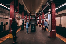 A subway platform with several people standing and engaging with their phones. The red columns and staircases are prominent features, along with signs indicating it's the Bowery station. The lighting is somewhat dim, characteristic of underground stations.