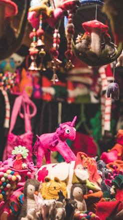 A vibrant display of colorful felt toys and decorations. A prominent pink unicorn stands out amidst a collection of various plush animals and ornaments, including penguins, mushrooms, and bells. The scene is filled with a whimsical assortment of handcrafted figures in bright hues of pink, red, and green.