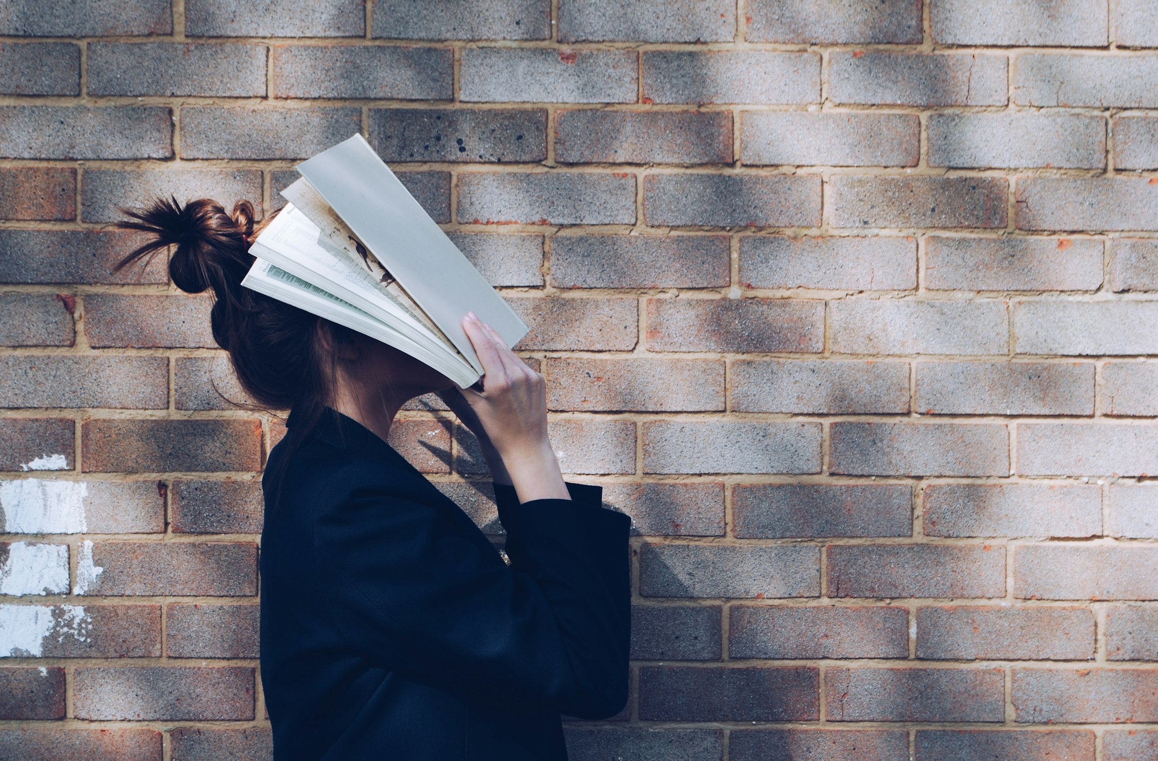 Girl stood next to a brick wall, holding a book over her face.