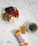 A small plate contains various snacks including kalamata olives, slices of pepperoni, blocks of cheese, and some seed crackers. Nearby, a round dish holds a few blueberries. A peanut butter protein bar, partially unwrapped, lies next to its packaging on a marble surface.