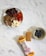 A small plate contains various snacks including kalamata olives, slices of pepperoni, blocks of cheese, and some seed crackers. Nearby, a round dish holds a few blueberries. A peanut butter protein bar, partially unwrapped, lies next to its packaging on a marble surface.