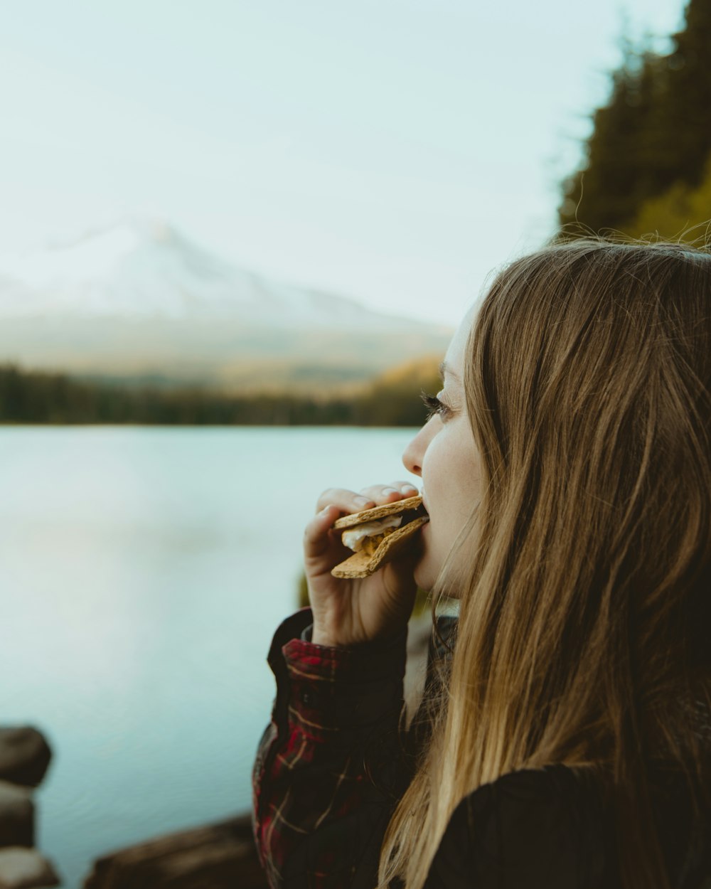 mujer de pie y comiendo alimentos frente al cuerpo de agua que ve la montaña