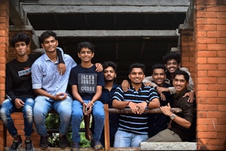 A group of eight young men are gathered closely together, posing cheerfully on a brick structure. They are casually dressed, with a mix of shirts and t-shirts, some wearing jeans. The atmosphere seems relaxed and friendly as they lean on a metal railing.
