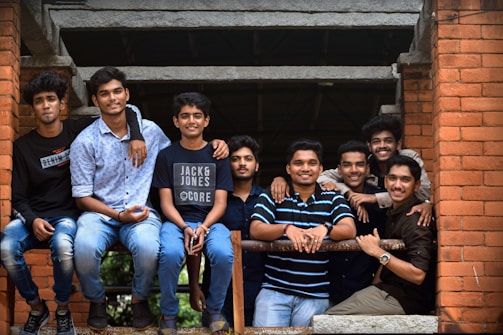 A group of eight young men are gathered closely together, posing cheerfully on a brick structure. They are casually dressed, with a mix of shirts and t-shirts, some wearing jeans. The atmosphere seems relaxed and friendly as they lean on a metal railing.