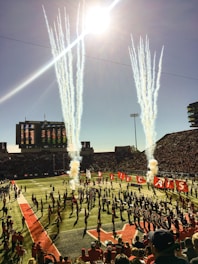 A lively football stadium scene with bright sunlight streaming from above. Large columns of smoke rise from the field, creating a dramatic effect. A marching band and cheerleaders perform on the field, carrying flags spelling out 'Wildcats'. The stands are filled with spectators, adding to the energetic atmosphere.
