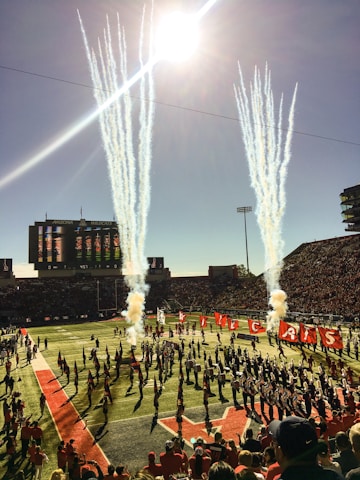 A lively football stadium scene with bright sunlight streaming from above. Large columns of smoke rise from the field, creating a dramatic effect. A marching band and cheerleaders perform on the field, carrying flags spelling out 'Wildcats'. The stands are filled with spectators, adding to the energetic atmosphere.