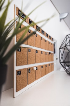 A wall-mounted cork board calendar with black and white day labels at the top, individual cork squares for each day, and white pushpins. The calendar is positioned at an angle, with a blurred plant in the foreground on the left and a geometric metal object on the right.