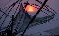 Large fishing nets are silhouetted against a vivid sunset, with the sun appearing as an orange orb in the sky. The nets are intricately woven and supported by a network of wooden beams. A small bird can be seen flying in the background, and there is a faint outline of a boat on the water.