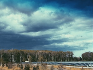 Dark, dramatic clouds loom over a field of solar panels set against a forest backdrop. The scene suggests an impending storm, with the vibrant greenery of the trees contrasting against the moody sky.