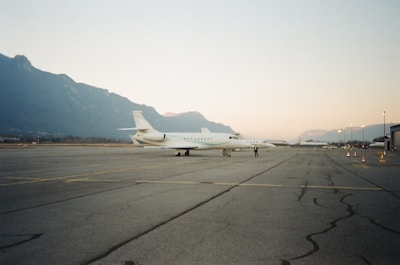 A private jet is parked on a vast, empty airport runway with mountainous terrain in the background under a clear sky. The runway is marked with yellow lines, and there are several orange traffic cones positioned along the side. A person is standing near the jet, adding a sense of scale to the scene.