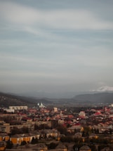 A view of a cityscape with various buildings, both residential and commercial, amidst a valley. The rooftops are mostly red and some buildings are yellow. In the background, there is a mountain range with snow-covered peaks under a partly cloudy sky.