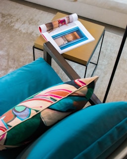 A cozy setting featuring a modern chair with a vibrant, abstract-patterned pillow. Nearby, a small wooden side table holds a colorful open magazine displaying interior designs and a folded magazine with a red cover. The setup rests on a textured, beige carpet, creating a warm, inviting atmosphere.