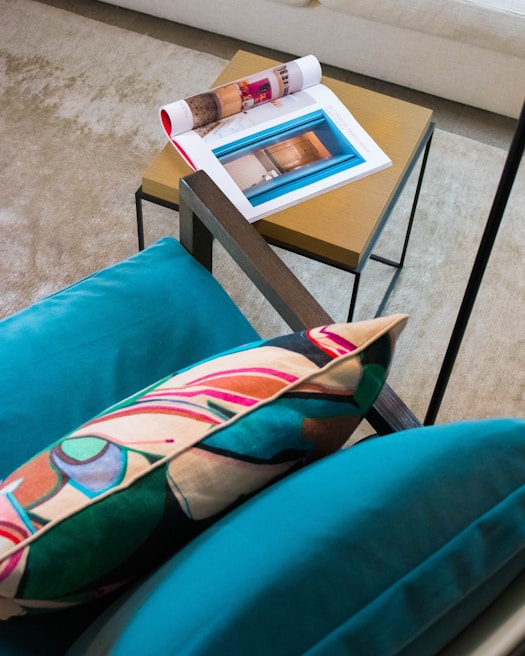 A cozy setting featuring a modern chair with a vibrant, abstract-patterned pillow. Nearby, a small wooden side table holds a colorful open magazine displaying interior designs and a folded magazine with a red cover. The setup rests on a textured, beige carpet, creating a warm, inviting atmosphere.