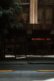 The image features an urban scene with a restaurant and bar. A neon sign with the words 'RESTAURANTE BAR' is visible against the backdrop of a building with glass and steel architecture. The street in front is dimly lit, creating a moody ambiance. Trees partially shade the area, adding a natural element to the urban setting.