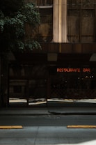 The image features an urban scene with a restaurant and bar. A neon sign with the words 'RESTAURANTE BAR' is visible against the backdrop of a building with glass and steel architecture. The street in front is dimly lit, creating a moody ambiance. Trees partially shade the area, adding a natural element to the urban setting.