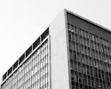 A black and white photograph of a modern, multi-story building. The structure has a grid of large windows covering its facade, indicating an office or commercial usage. The composition focuses on the sharp lines and geometric forms of the building.