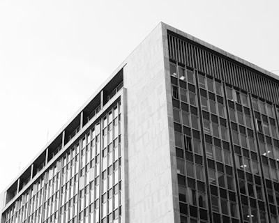 A black and white photograph of a modern, multi-story building. The structure has a grid of large windows covering its facade, indicating an office or commercial usage. The composition focuses on the sharp lines and geometric forms of the building.