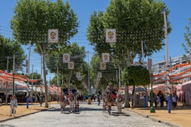A festive street scene with vibrant colors, featuring horse-drawn carriages and people dressed in traditional attire. Striped tents line both sides of the paved road, adorned with colorful bunting and lights. Tall, leafy trees provide shade along the street, adding to the lively atmosphere.