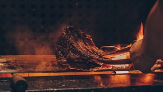 A large piece of steak being grilled over an open flame with tongs holding the meat. The image has a warm, smoky atmosphere with visible flames and a dark, rustic background.