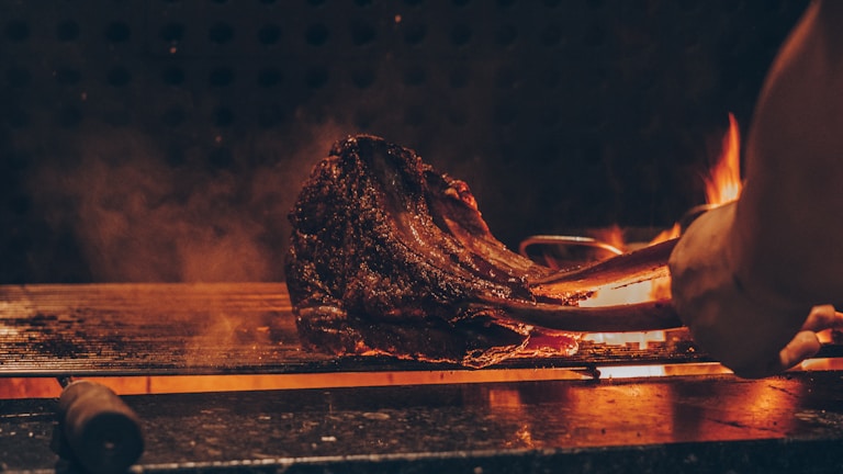 Rounded photo of a sizzling steak on the grill with flames around.