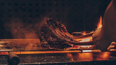 A large piece of steak being grilled over an open flame with tongs holding the meat. The image has a warm, smoky atmosphere with visible flames and a dark, rustic background.