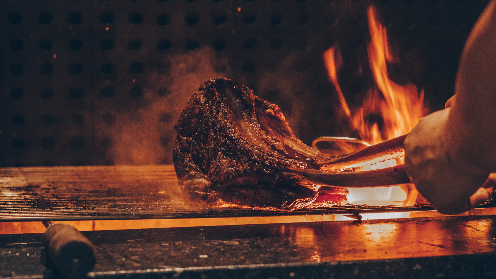 A thick, charred steak seared over glowing open-fire embers in a dark kitchen