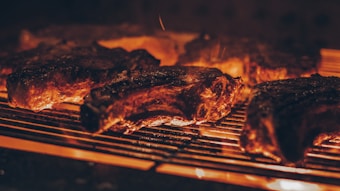 Several pieces of meat are being grilled over an open flame. The surface of the meat is charred, indicating high heat and searing. The glowing embers and fiery orange hue create a warm atmosphere.