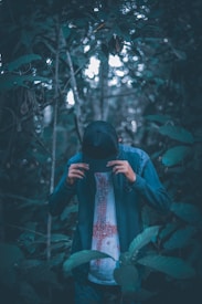 A person wearing a dark baseball cap and a jacket stands amidst dense foliage in a forest setting. The individual holds the cap with both hands, with their head slightly bowed. The background is filled with blurred leaves and trees, creating a mysterious and moody ambiance.