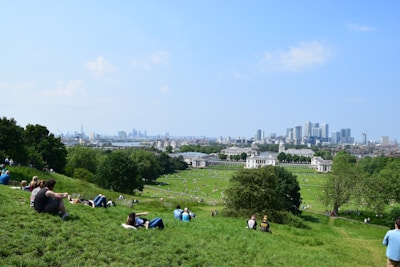 A green park with people relaxing and lying on the grass. The park is situated near a historic building complex with a city skyline in the background under a clear blue sky.