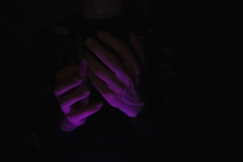 Hands illuminated by a soft purple light emerge from a dark background, creating a sense of mystery and focus on the illuminated area.