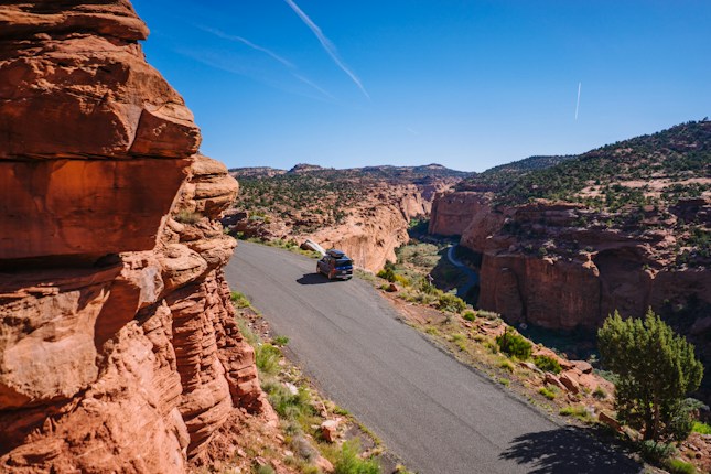 A car is traveling on a narrow road that winds through a desert landscape characterized by red rock formations and deep canyons. The sky is clear blue, and the sun casts shadows highlighting the textures of the rocks. Sparse green vegetation can be seen along the road and cliffs.