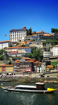 A scenic view of a hillside town featuring colorful buildings with red-tiled roofs. A river runs in front, with a traditional boat filled with passengers cruising along. The sky is clear and blue, adding a vibrant backdrop to the lush greenery scattered across the landscape.