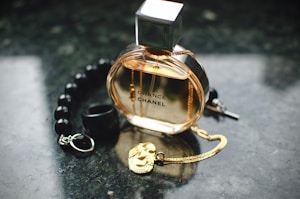 A glass perfume bottle labeled 'Chance Chanel' is placed on a dark, reflective surface. Surrounding it are a string of black beads, a small black ring, a gold necklace with a pendant, and a black pen cap.