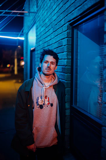 A moody street scene with a glowing neon sign reflecting off a charcoal wall and a model wearing a 3D-rendered hoodie.