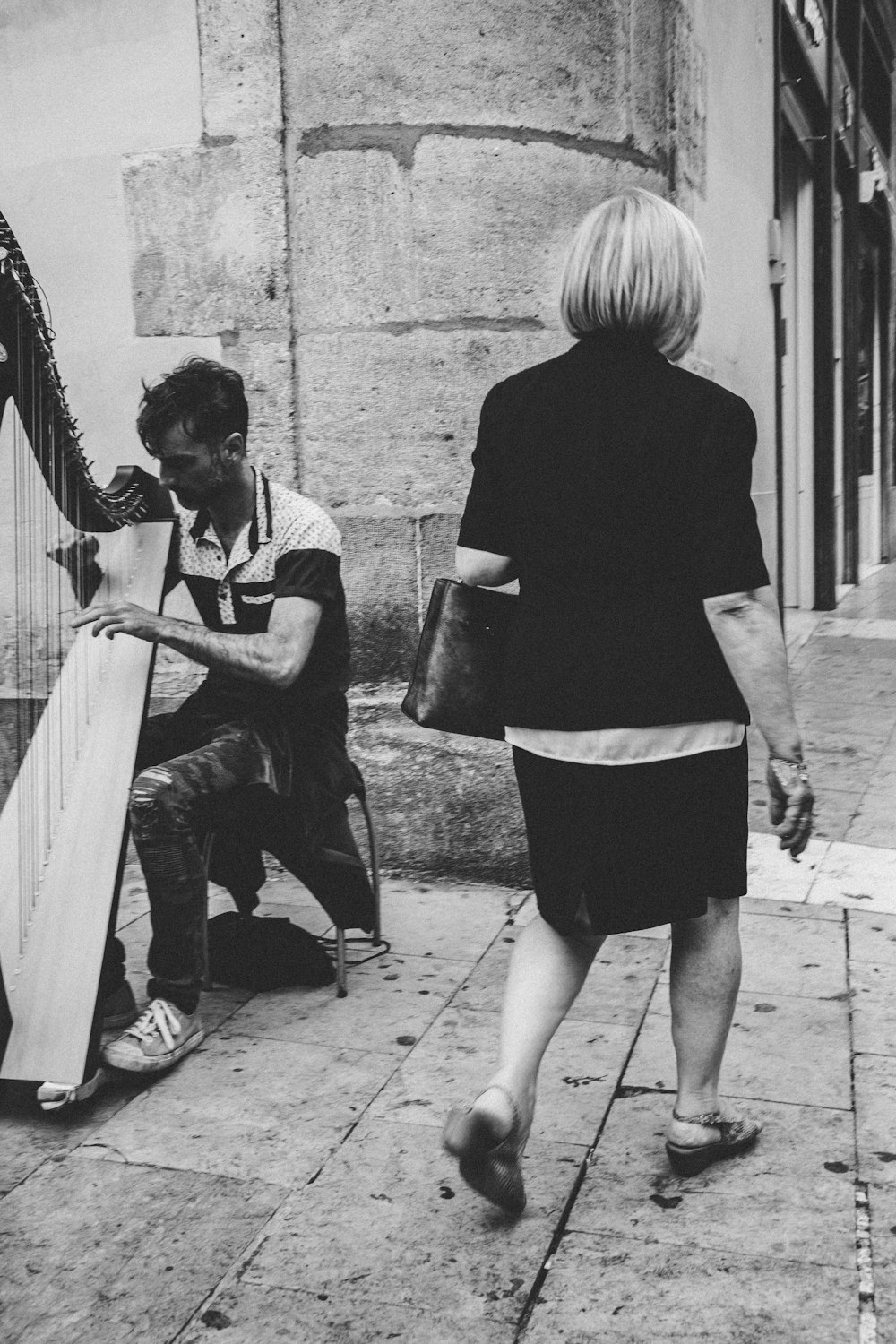 grayscale photo of woman walking beside man playing harp