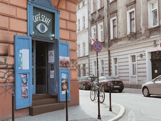 A quaint street corner with a café featuring a blue door and sign with the name 'Café Szafé'. Posters adorn the doorway and walls, and a bicycle is parked on the sidewalk. Several cars are visible on the street, along with a traffic sign indicating no parking.