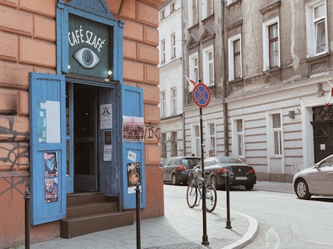 A quaint street corner with a café featuring a blue door and sign with the name 'Café Szafé'. Posters adorn the doorway and walls, and a bicycle is parked on the sidewalk. Several cars are visible on the street, along with a traffic sign indicating no parking.