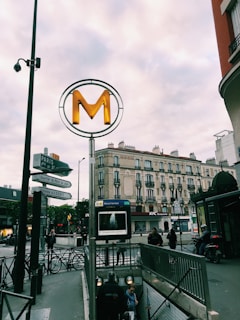 A Parisian metro entrance with a large 'M' sign is set against a background of classic Parisian architecture. People are descending the stairs to the metro, and various street signs are visible. A KFC restaurant is in the background along with several motorbikes parked nearby. The sky is overcast, adding a subdued tone to the scene.