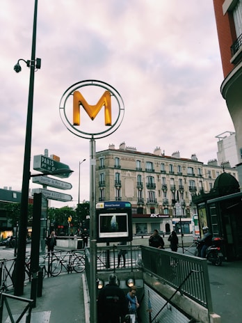 A Parisian metro entrance with a large 'M' sign is set against a background of classic Parisian architecture. People are descending the stairs to the metro, and various street signs are visible. A KFC restaurant is in the background along with several motorbikes parked nearby. The sky is overcast, adding a subdued tone to the scene.