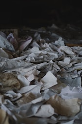 A chaotic pile of crumpled and torn paper scattered on what appears to be a floor, with dim lighting contributing to a somber atmosphere.