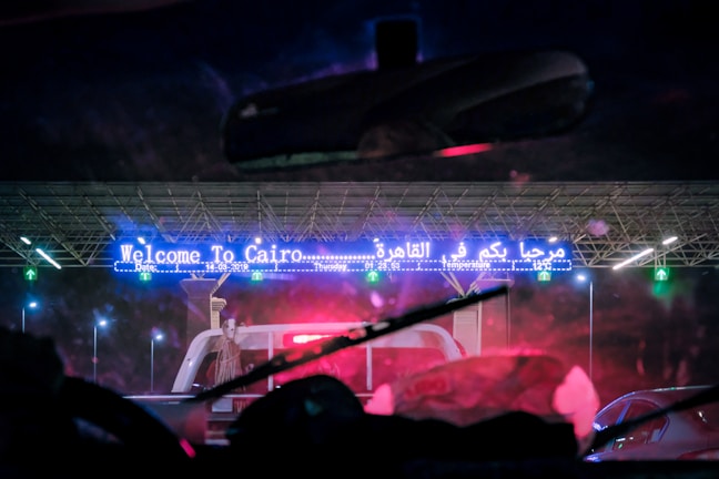 A nighttime scene through a vehicle's windshield shows a large illuminated sign reading 'Welcome To Cairo' in both English and Arabic. There are multiple lanes and vehicles passing through, with a toll booth visible. Bright lights illuminate the area, highlighting the structure's modern design.